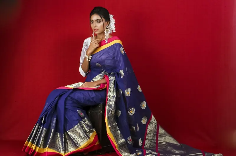 Woman in a blue silk saree seated low with the pallu spread across the floor