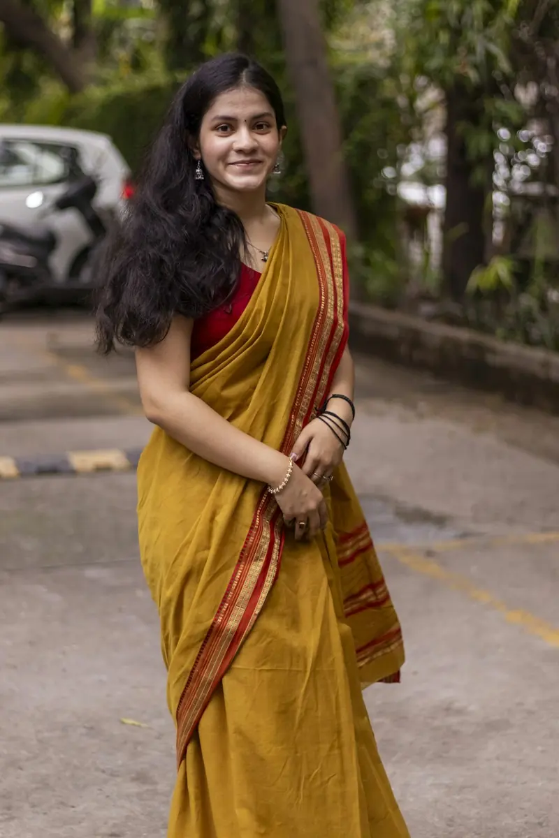 Woman in a mustard cotton saree standing on a street, holding the pallu and smiling