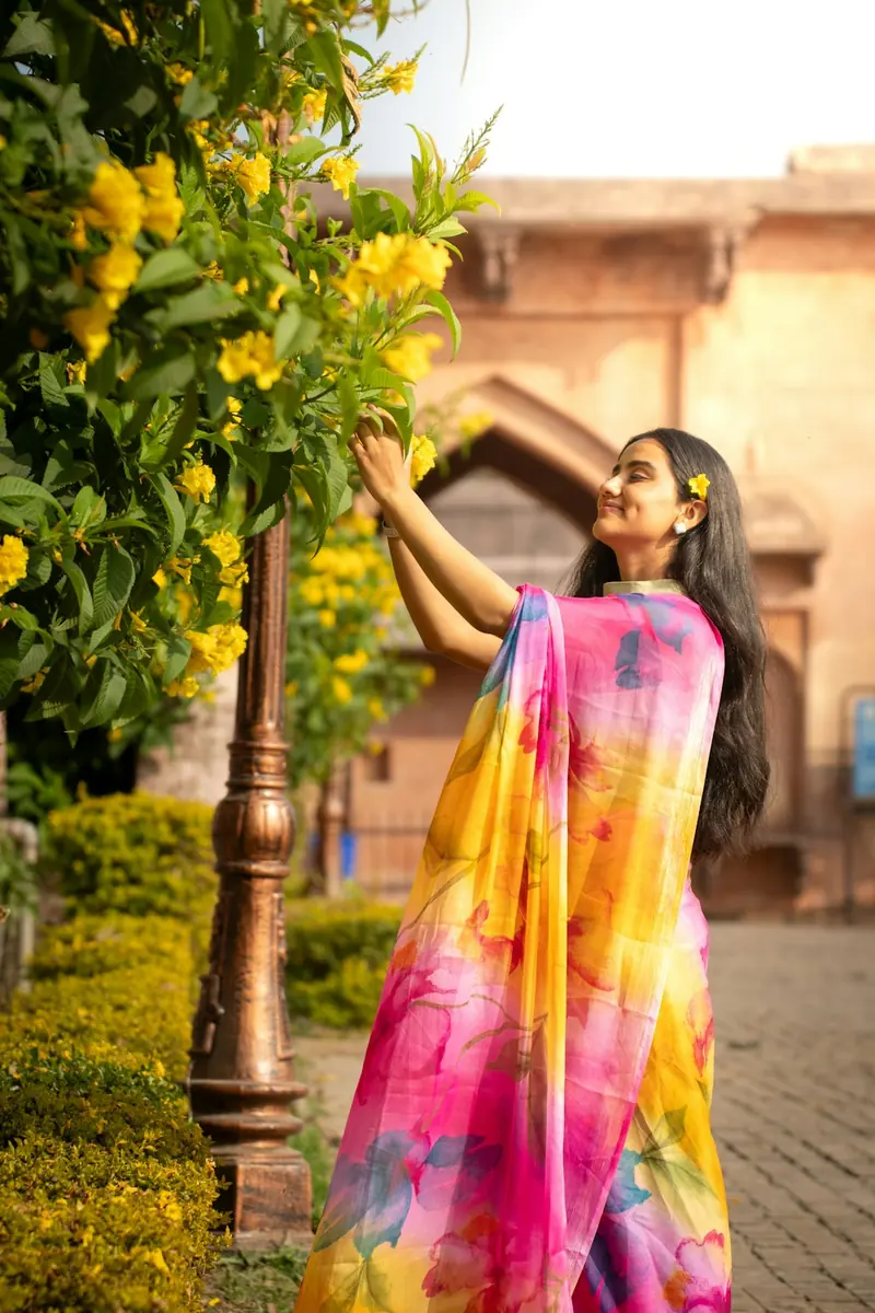 Woman in a colourful saree reaching up towards yellow flowers on a tree
