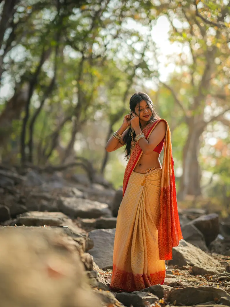 Woman in an orange and cream saree adjusting her earrings with both hands in a forest