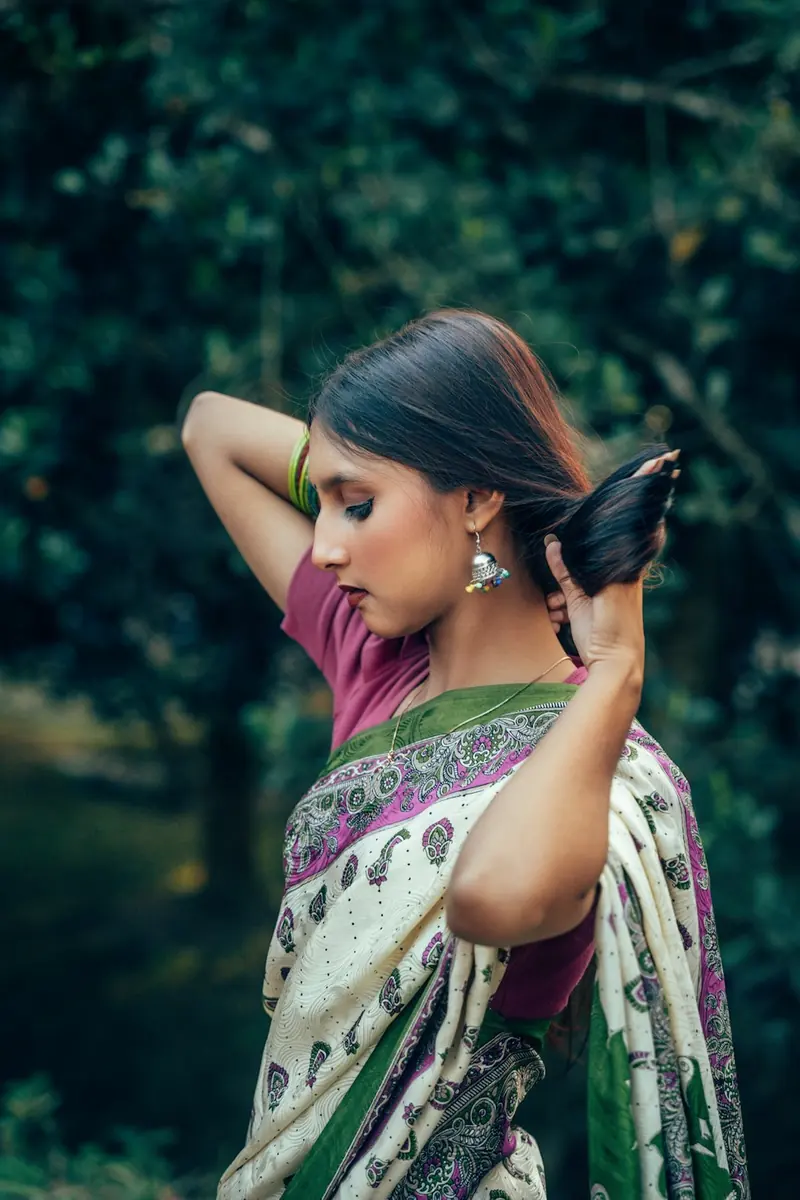 Woman in a green and white saree with both hands in her hair, looking down
