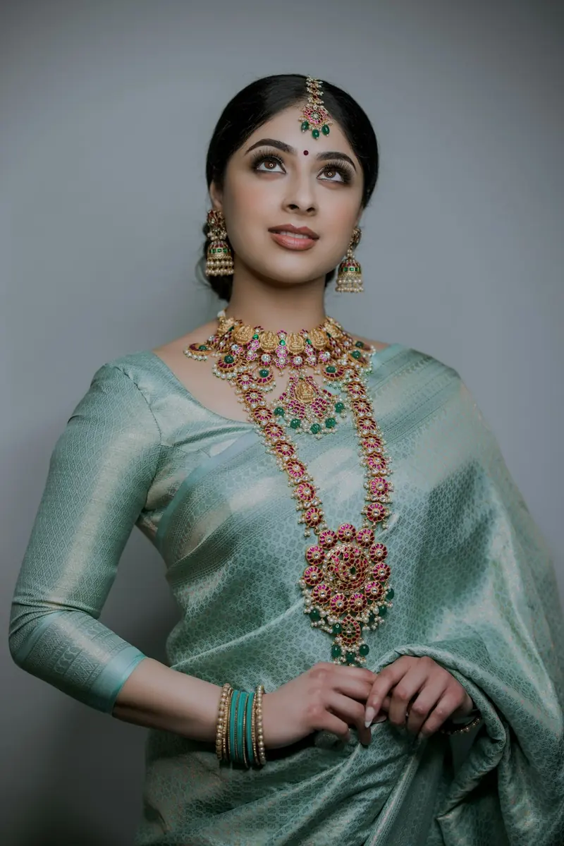 Woman in a green silk saree with a long temple jewellery necklace, hands clasped and looking up