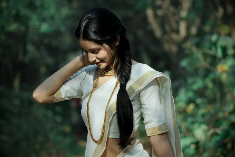 Woman in a white and gold Kerala saree with a long braid over her shoulder, looking down