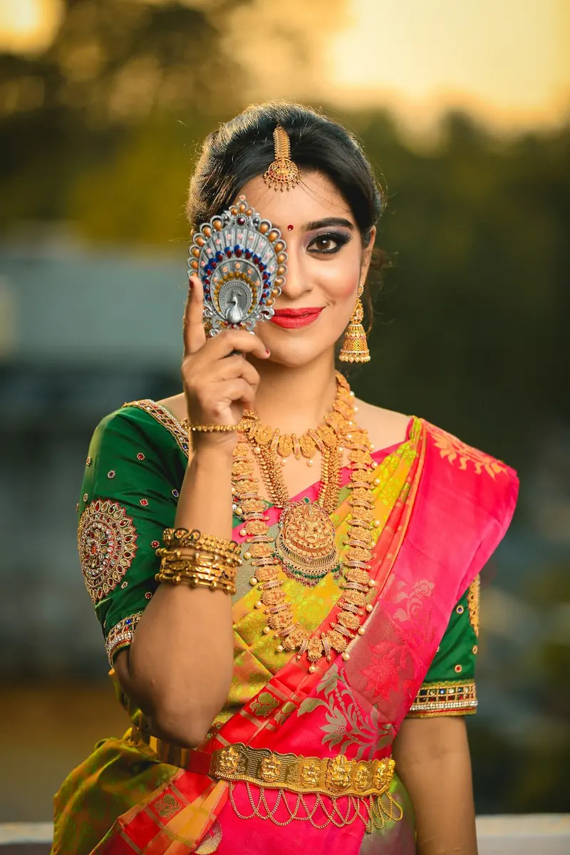 Woman in a pink and green silk saree covering one eye with a decorative peacock hand fan