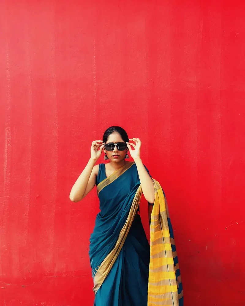 Woman in a blue and yellow saree adjusting sunglasses against a red wall