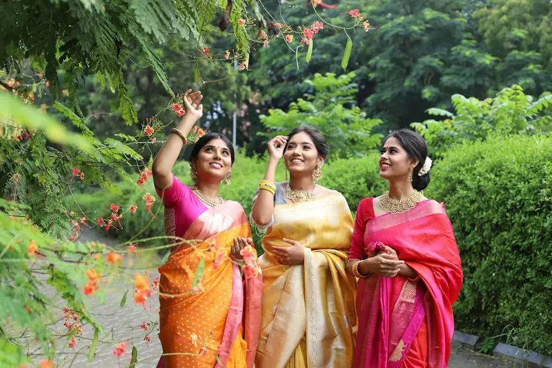 Three women in bright silk sarees laughing together in a garden