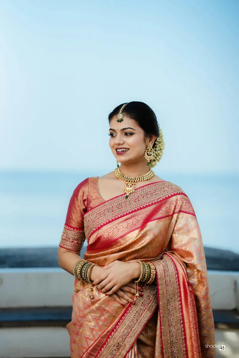 Woman in a red and gold silk saree with hands clasped, smiling and looking to the side