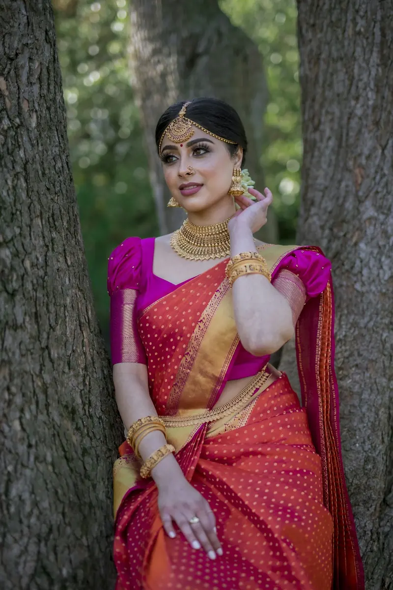 Woman in a pink and gold silk saree standing between two tree trunks, touching her earring