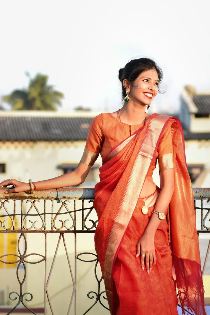 Woman in an orange saree leaning on a balcony railing and smiling