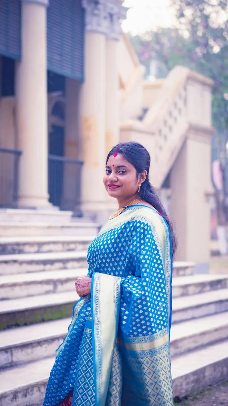 Woman in a blue Banarasi saree standing at the foot of stone steps, smiling at the camera
